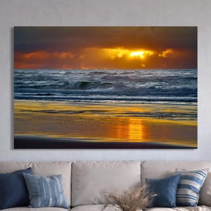 May include: A rectangular wall art print featuring a sunset over the ocean. The scene displays dark clouds with a bright golden sun glowing behind them, casting a vibrant yellow reflection across the wet sand and the rolling blue waves of the sea.