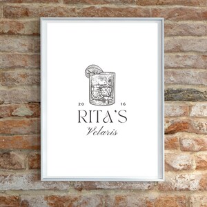 May include: A white framed print with a black and white illustration of a cocktail glass with a lemon wedge. The text "Rita's Velaris" is below the illustration.