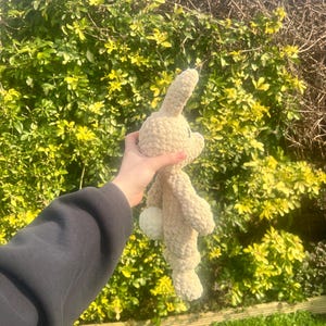 May include: A crocheted beige bunny rabbit plush toy with a white fluffy tail. The toy is being held in a person's hand.