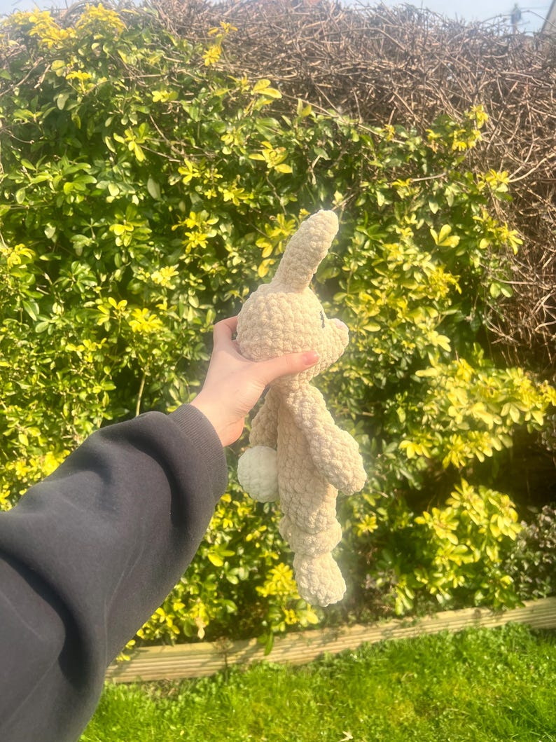 May include: A crocheted beige bunny rabbit plush toy with a white fluffy tail. The toy is being held in a person's hand.