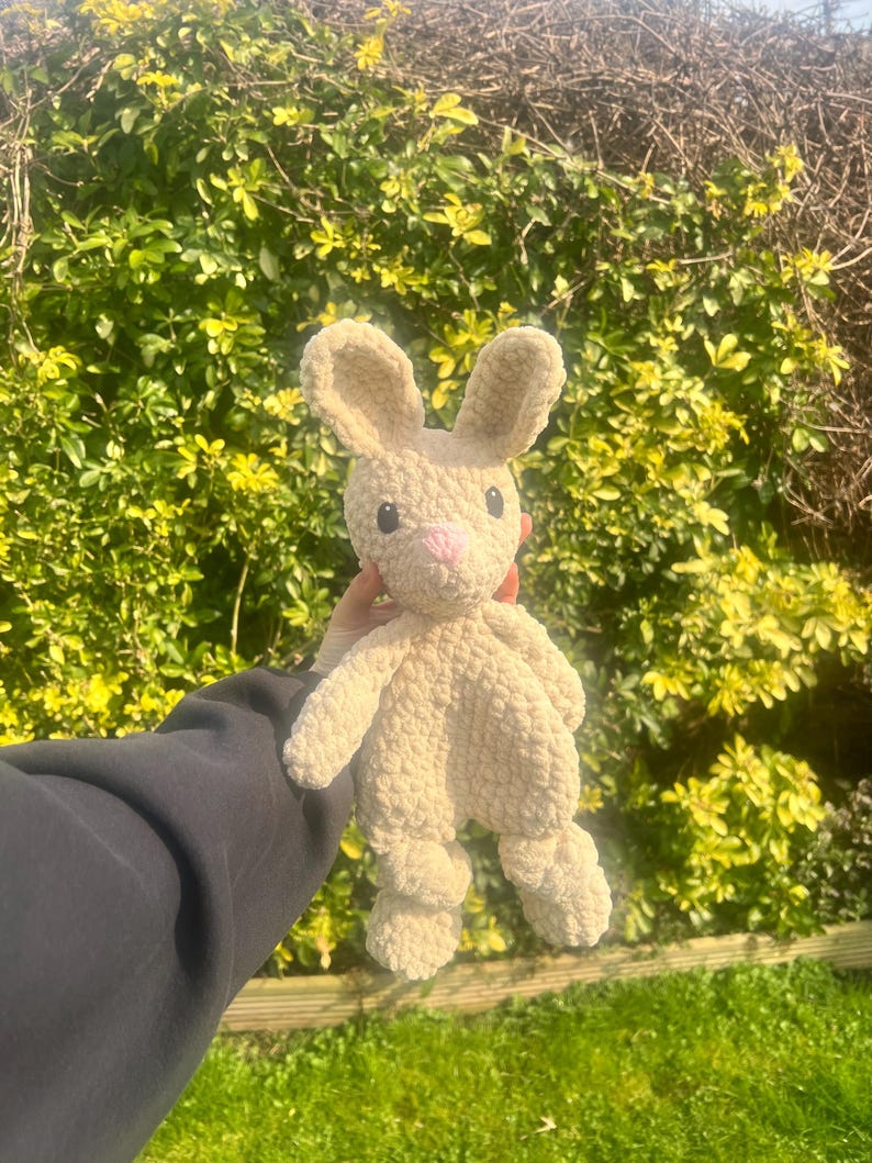 May include: A crocheted plush bunny toy in a cream color with pink nose and eyes. The bunny is being held in front of a green bush.