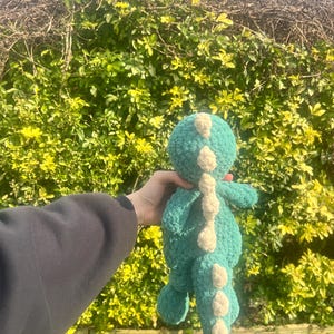 May include: A teal green crocheted dinosaur toy with white accents. The dinosaur has a long tail and a small head with a white spot on top.
