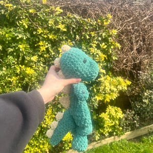 May include: A green crocheted dinosaur toy with white accents. The dinosaur is being held by a hand.