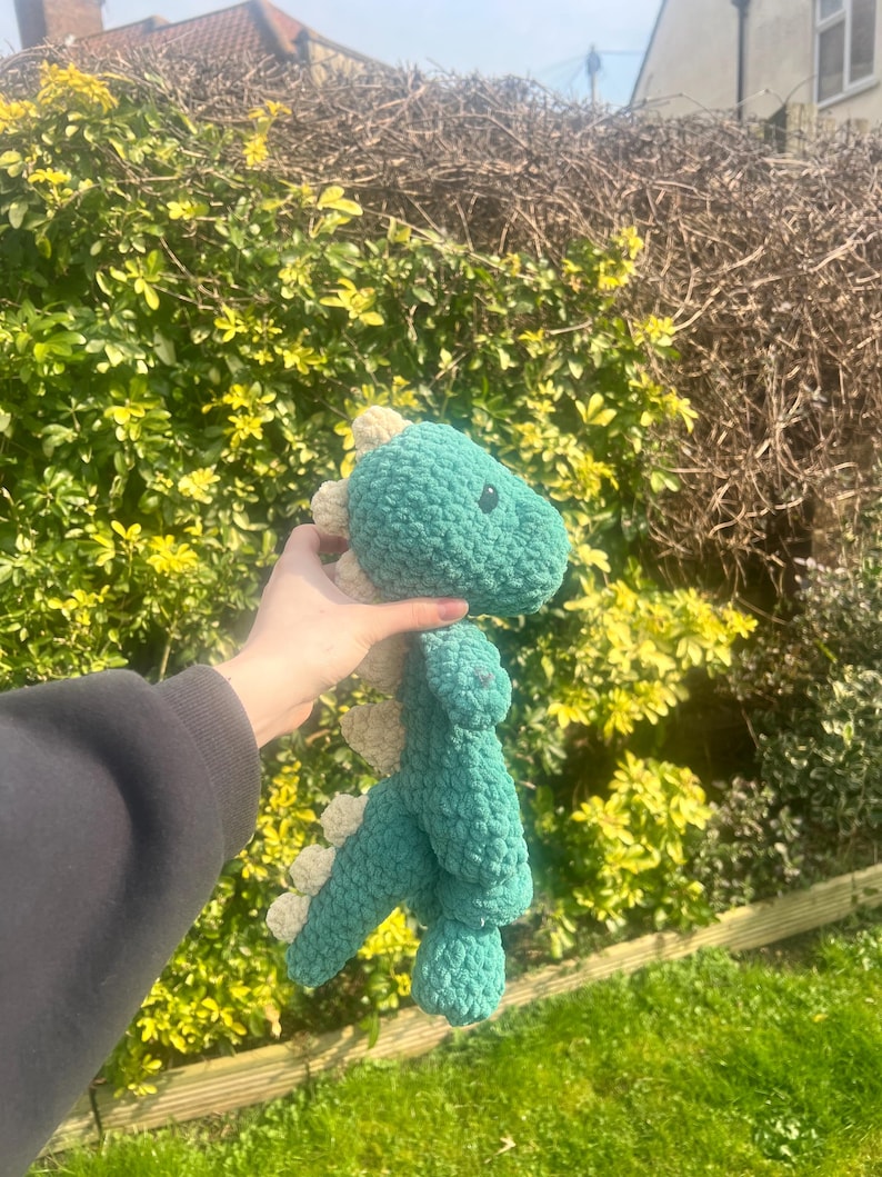 May include: A green crocheted dinosaur toy with white accents. The dinosaur is being held by a hand.