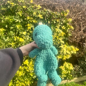 May include: A green crocheted dinosaur plush toy with a small white horn on its head. The toy is being held in a person's hand.