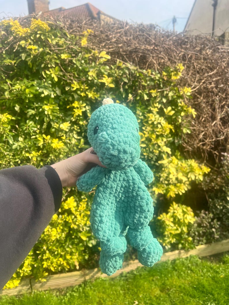 May include: A green crocheted dinosaur plush toy with a small white horn on its head. The toy is being held in a person's hand.