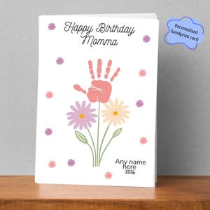 May include: A white greeting card featuring the text Happy Birthday Momma at the top and Any name here 2026 at the bottom. The central design shows a pink handprint serving as a flower head atop green stems with purple and yellow daisies.