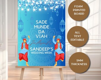 Op de afbeelding: Een blauw en turquoise trouwteken op een ezel. Het teken bevat de tekst "SADE MUNDE DA VIAH" en "SANDEEP'S WEDDING WEEK". Het bevat illustraties van figuren in traditionele kledij en decoratieve elementen.