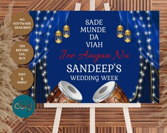 Op de afbeelding: Een marineblauw trouwbord op een witte ezel. Het bord heeft tekst in wit en rood, met gouden lantaarns en een drumafbeelding. Op het bord staat "Sandeep's Wedding Week".