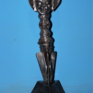 May include: A silver-coloured metal statue of a deity with a fierce expression, holding a dagger. The statue is mounted on a black, triangular base.