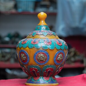 Puede incluir: Una urna decorativa amarilla, azul y roja con diseños intrincados y una parte superior redonda. La urna está hecha de un mosaico de pequeñas piedras y tiene una base con un diseño de flor de loto.