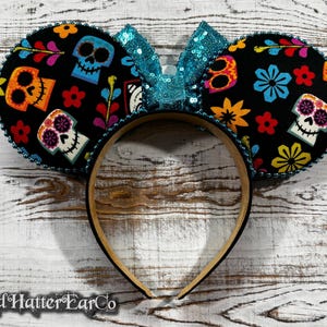 May include: Black mouse ear headband featuring colorful sugar skull designs and a turquoise sequin bow. The ears are decorated with flowers and other festive elements. The headband is trimmed with a beaded edge.