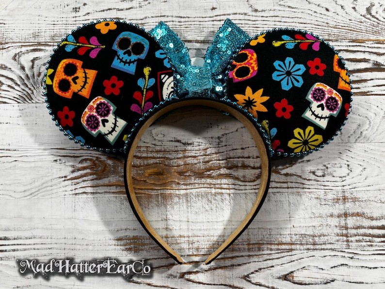 May include: Black mouse ear headband featuring colorful sugar skull designs and a turquoise sequin bow. The ears are decorated with flowers and other festive elements. The headband is trimmed with a beaded edge.