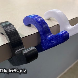 May include: Three 3D printed hooks in black, blue, and white. The hooks are designed to hang over the edge of a table or shelf.
