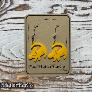 May include: A pair of yellow dangle earrings featuring a silhouette of Simba from the Lion King movie. The earrings are on a brown card with the text "Mad Hatter Ear Co."