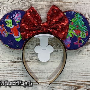 May include: A festive headband featuring Grinch-themed ears and a sequined red bow. The ears display Grinch characters with gifts and a Christmas tree on a navy blue background. The headband has a white Mickey Mouse silhouette.