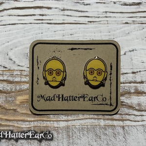 May include: A pair of gold and yellow C-3PO earrings on a brown card with the text "Mad Hatter Ear Co."