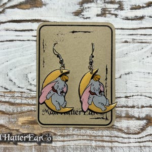 May include: A pair of dangle earrings featuring a yellow crescent moon with a pink and gray Dumbo the Elephant illustration. The earrings are on a brown card with the text "Mad Hatter EarCo".
