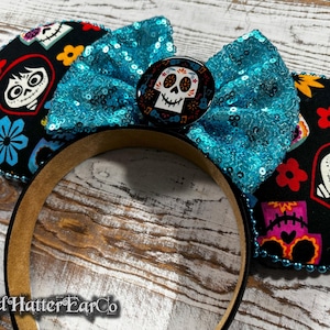 May include: A black headband with Coco-themed ears. The ears feature colorful floral and skull designs, with a turquoise sequined bow and a center button. The headband is trimmed with blue beads.