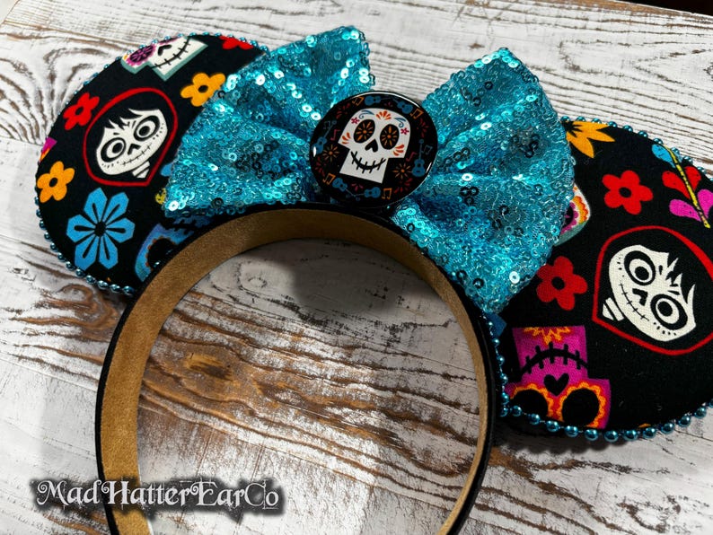 May include: A black headband with Coco-themed ears. The ears feature colorful floral and skull designs, with a turquoise sequined bow and a center button. The headband is trimmed with blue beads.