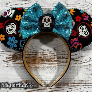 May include: A headband with black ears featuring colorful floral and character designs. A turquoise sequined bow sits between the ears, with a skull-themed center. The headband is brown and black, with the text "Mad Hatter EarCo" at the bottom.