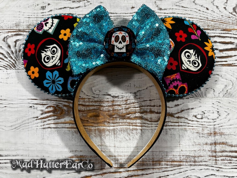 May include: A headband with black ears featuring colorful floral and character designs. A turquoise sequined bow sits between the ears, with a skull-themed center. The headband is brown and black, with the text "Mad Hatter EarCo" at the bottom.