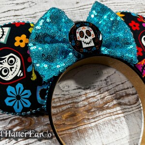 May include: Black mouse ears with a turquoise sequined bow and a Day of the Dead skull button. The ears are decorated with colorful floral and skull patterns. The headband is brown.