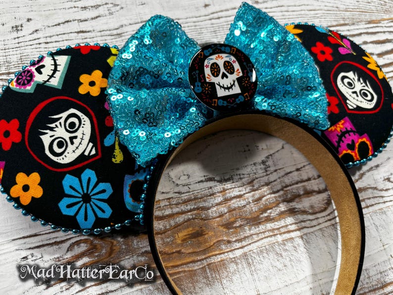 May include: Black mouse ears with a turquoise sequined bow and a Day of the Dead skull button. The ears are decorated with colorful floral and skull patterns. The headband is brown.
