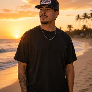 Puede incluir: Gorra de b&eacute;isbol negra con visera plana y un parche rectangular con la palabra "Merica". La persona lleva una camiseta negra y una cadena de plata. El fondo es una playa al atardecer.
