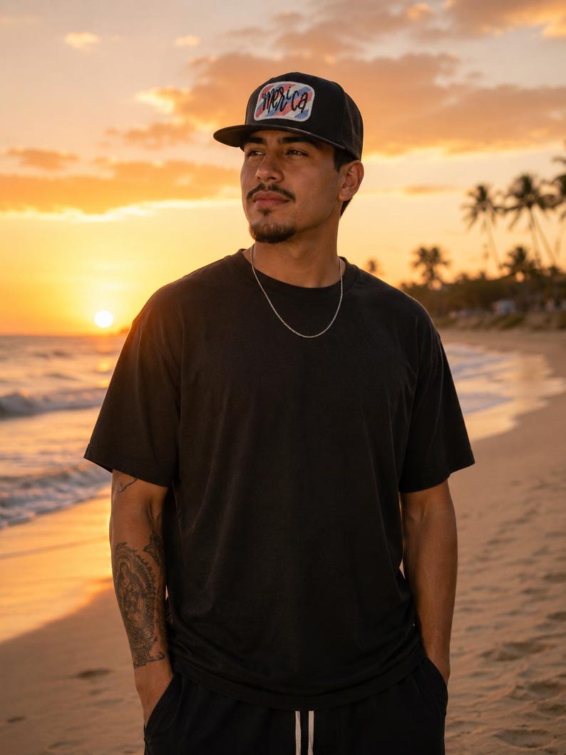 Puede incluir: Gorra de b&eacute;isbol negra con visera plana y un parche rectangular con la palabra "Merica". La persona lleva una camiseta negra y una cadena de plata. El fondo es una playa al atardecer.