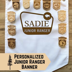 May include: A white fabric banner with the text "Personalized Junior Ranger Banner" and a brown wooden tree silhouette. The banner has a brown border and is decorated with various wooden Junior Ranger badges from different national parks, including Voyageurs, Tonto, Tuzigoot, Casa Grande Ruins, Wupatki, Walnut Canyon, Montezuma Castle, Sunset Crater Volcano, South Rim, and Saguaro.