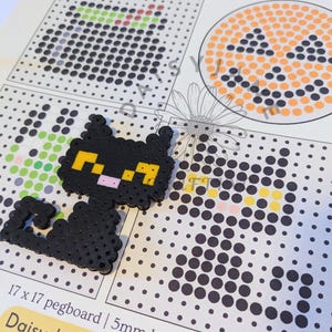 May include: A black cat figure made of fused plastic beads with yellow eyes and a pink nose, placed on a printed paper Halloween perler bead template. The template shows a 17 x 17 pegboard grid and specifies 5mm beads for the crafting project.