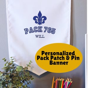 May include: White banner with a blue fleur-de-lis and the text "PACK 765 WILL". The banner has two metal grommets at the top for hanging. An oval graphic below reads "Personalized Pack Patch & Pin Banner".