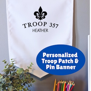 May include: White banner with a black fleur-de-lis emblem and text that reads "TROOP 357 HEATHER." The banner has metal grommets at the top for hanging. A blue oval graphic states "Personalized Troop Patch & Pin Banner."
