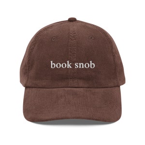 May include: A brown corduroy baseball cap with the text "book snob" embroidered in white on the front.