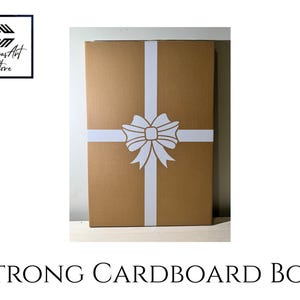 May include: A rectangular brown cardboard box featuring a white ribbon graphic printed in a cross pattern with a central bow. The box is labelled with the text Canvas Art Store in the top left corner and Strong Cardboard Box at the bottom.