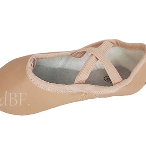 Puede incluir: Un par de zapatillas de ballet de cuero marrón claro con correas elásticas en la parte superior. Las zapatillas tienen un logotipo dBF en el lateral.