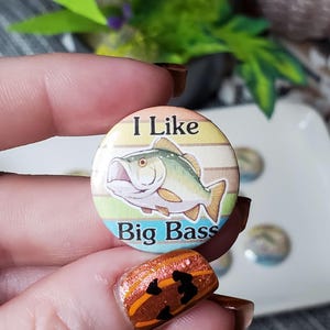 May include: A round button with the text "I Like Big Bass" and a graphic of a fish. The button has a striped background in shades of yellow, green, and blue. The button is held in a hand with brown nail polish.