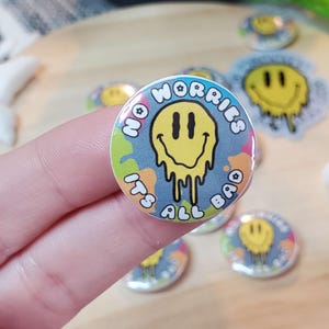 May include: A round button with the text "NO WORRIES ITS ALL BAD" encircling a yellow melting smiley face. The button has a light blue background with colorful paint splatters. Other buttons with the same design are visible.