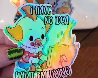 May include: A holographic sticker featuring a cartoon clown with a red nose and a blue and white outfit. The clown is sitting at a computer that is on fire. The text on the sticker reads "I have no idea what I'm doing".