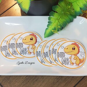 May include: A set of eight round stickers featuring a cartoon dinosaur with the text "Bite Me" in a yellow circle with a white background. The stickers are on a white surface.