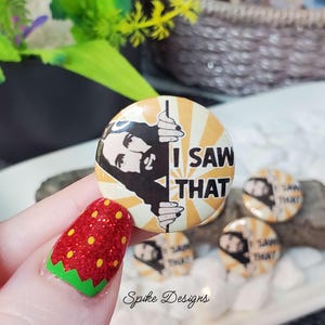 May include: A collection of round, yellow and white buttons with the text "I SAW THAT" and a black and white graphic. The buttons are held in a hand with a strawberry-themed nail design. The buttons are from Spike Designs.