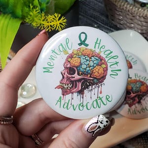 May include: A white button with a green ribbon and the text "Mental Health Advocate" in green lettering. A colorful illustration of a skull with a brain inside is in the center of the button.