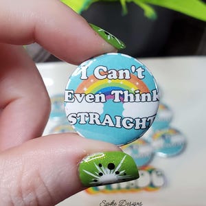 May include: A round button with a light blue background, a rainbow, and the text "I Can't Even Think STRAIGHT". The button is held between fingers with kiwi fruit nail art. Other buttons are visible in the background.