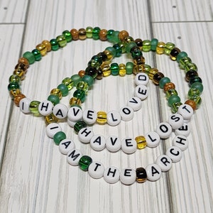 Op de afbeelding: Drie kralenarmbanden met groene, gouden en witte kralen. De armbanden spellen de zin "Ik heb liefgehad, ik ben de grootste".