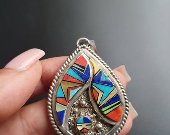 May include: A silver pendant with a teardrop shape, featuring a colorful inlay of turquoise, red, blue, and yellow stones. The pendant is set in a silver bezel and has a textured border.