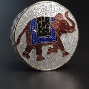 May include: A silver and enamel trinket box with a detailed image of an elephant in red, blue, and black enamel. The elephant is wearing a blue and black saddle.
