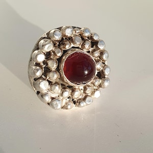 May include: A silver ring with a central, round, deep red gemstone. The gemstone is surrounded by a circle of small, silver, ball-shaped embellishments. The ring is set against a white background, with sunlight casting a shadow.