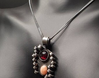 Puede incluir: Un collar de cadena de plata con un colgante geométrico que presenta dos piedras preciosas ovaladas. La piedra más grande es de color rojo intenso, mientras que la más pequeña es de color rosa pálido.