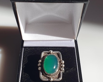 May include: A silver ring with a green oval gemstone set in a square bezel. The ring is surrounded by small silver beads.
